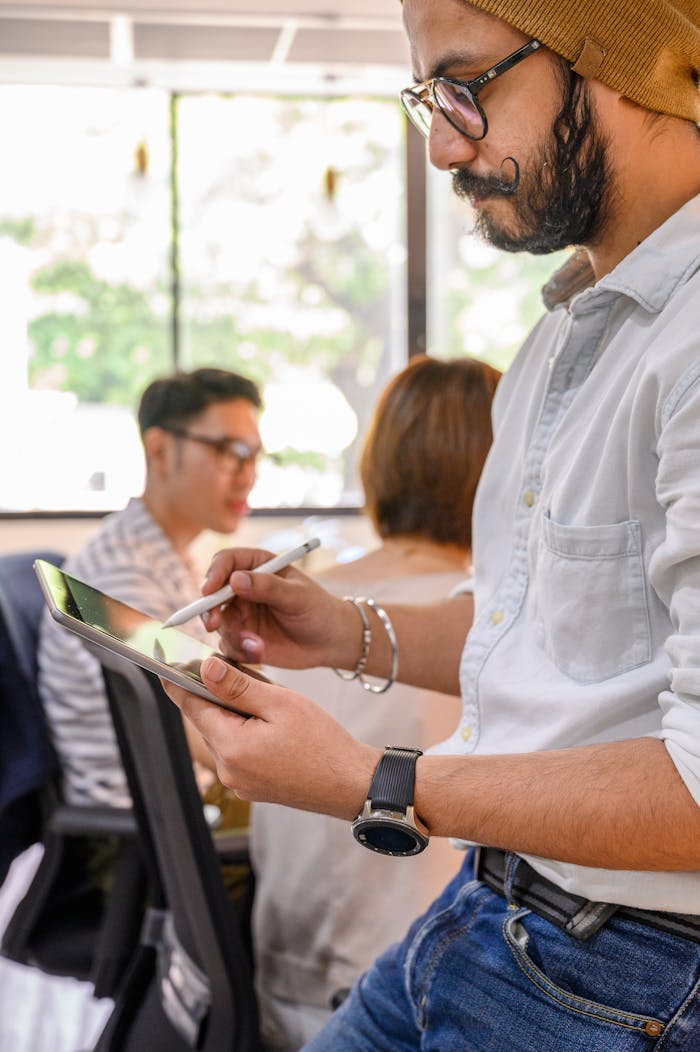 Bearded man wearing glasses and beanie, using tablet with stylus in a modern office.